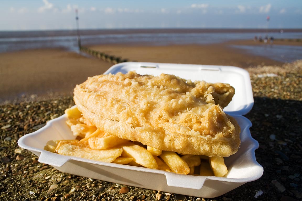 A thickly beer-battered fried fish fillet served with chips in a takeaway container