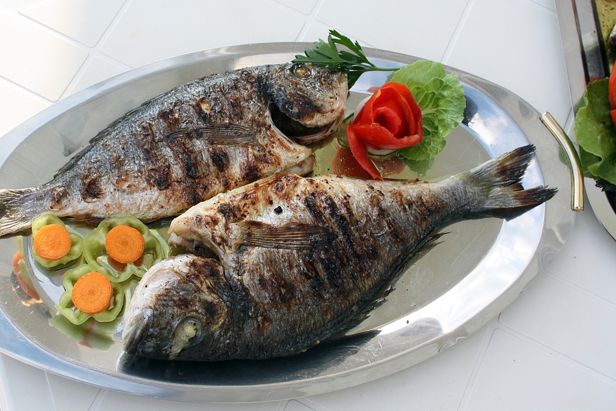 Two whole fish, scaled and gutted, grilled with visible char marks and garnished with lemon, carrot, and pepper